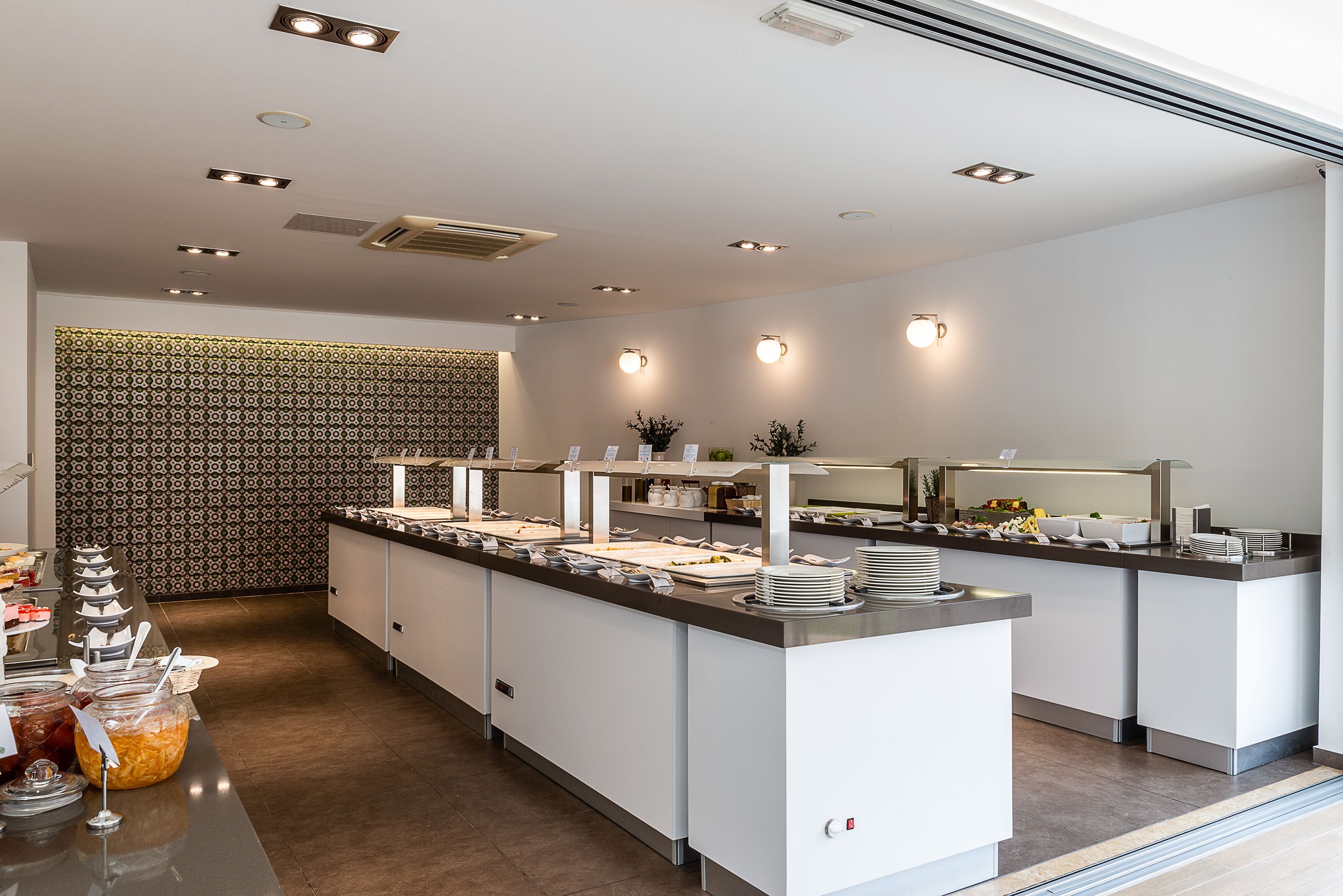 A modern buffet setup with a long counter, multiple serving stations, and a patterned wall in the background.