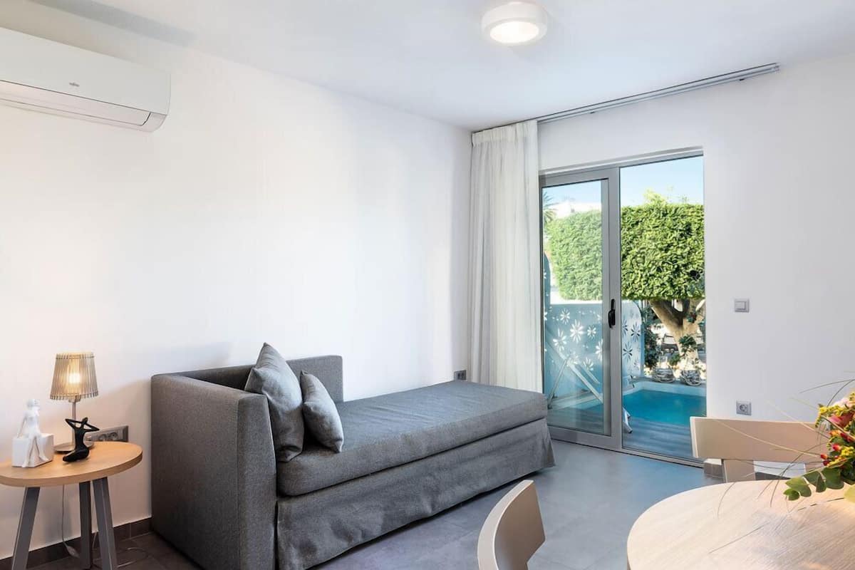 A modern room with a grey sofa, a small round table with a lamp, and a view of a swimming pool through glass doors.