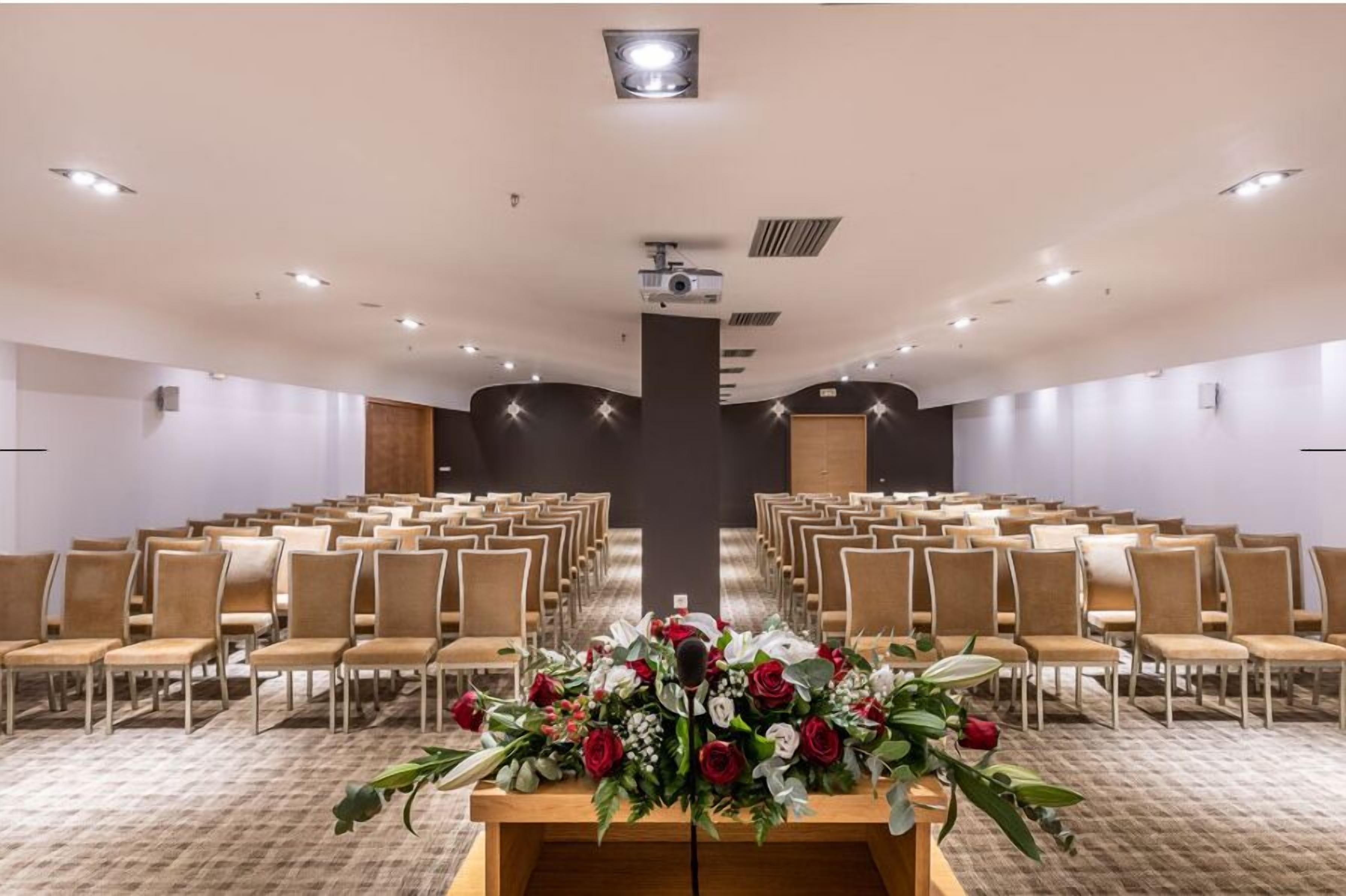 A conference room with rows of chairs, a podium with floral arrangements, and ceiling lights.