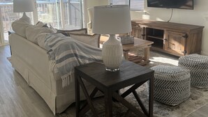 A living room with a sofa, a lamp, ottomans, a TV, and a view of the outdoors. - Large 4 bedroom penthouse unit 5 miles from Bethany Beach (Ocean View)