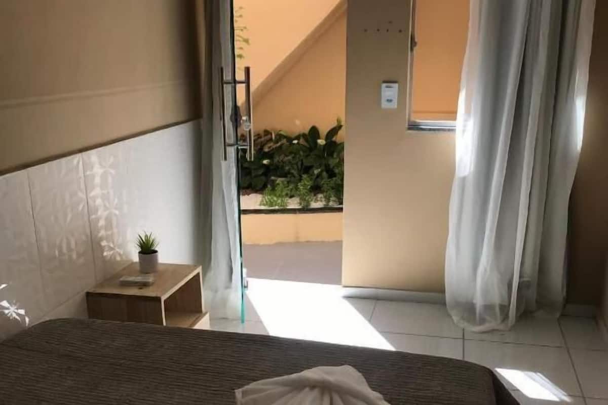 Une chambre à coucher avec un lit, une petite table de chevet, une plante en pot et une vue sur un balcon agrémenté de plantes et d’un escalier.