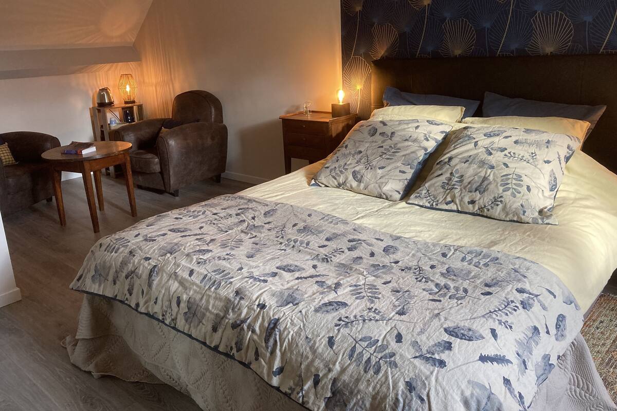 A bedroom with a large bed, a chair, a small table, and a nightstand with a lamp.
