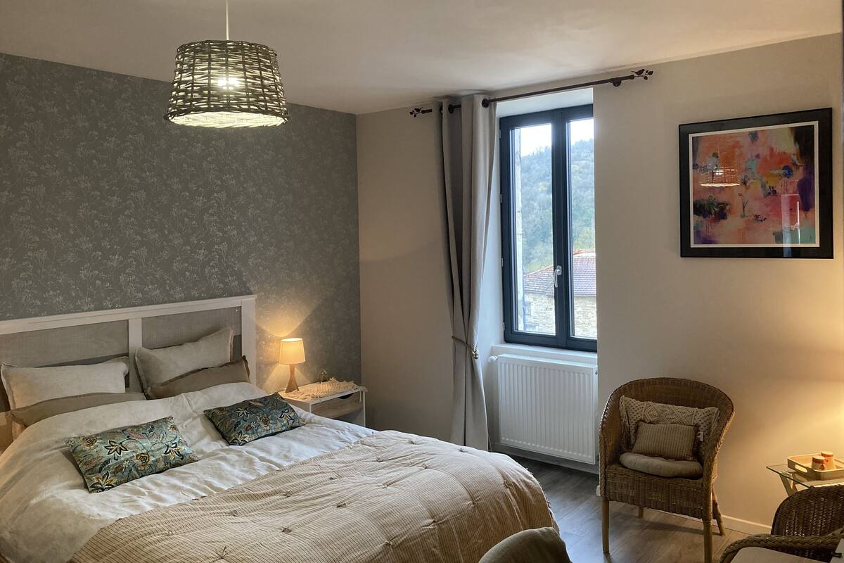 A bedroom with a bed, a bedside lamp, a wicker chair, a window with curtains, and a framed artwork on the wall.
