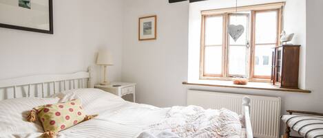A bedroom with a bed, a nightstand, a lamp, a window with a heart decoration, and a wooden chair.