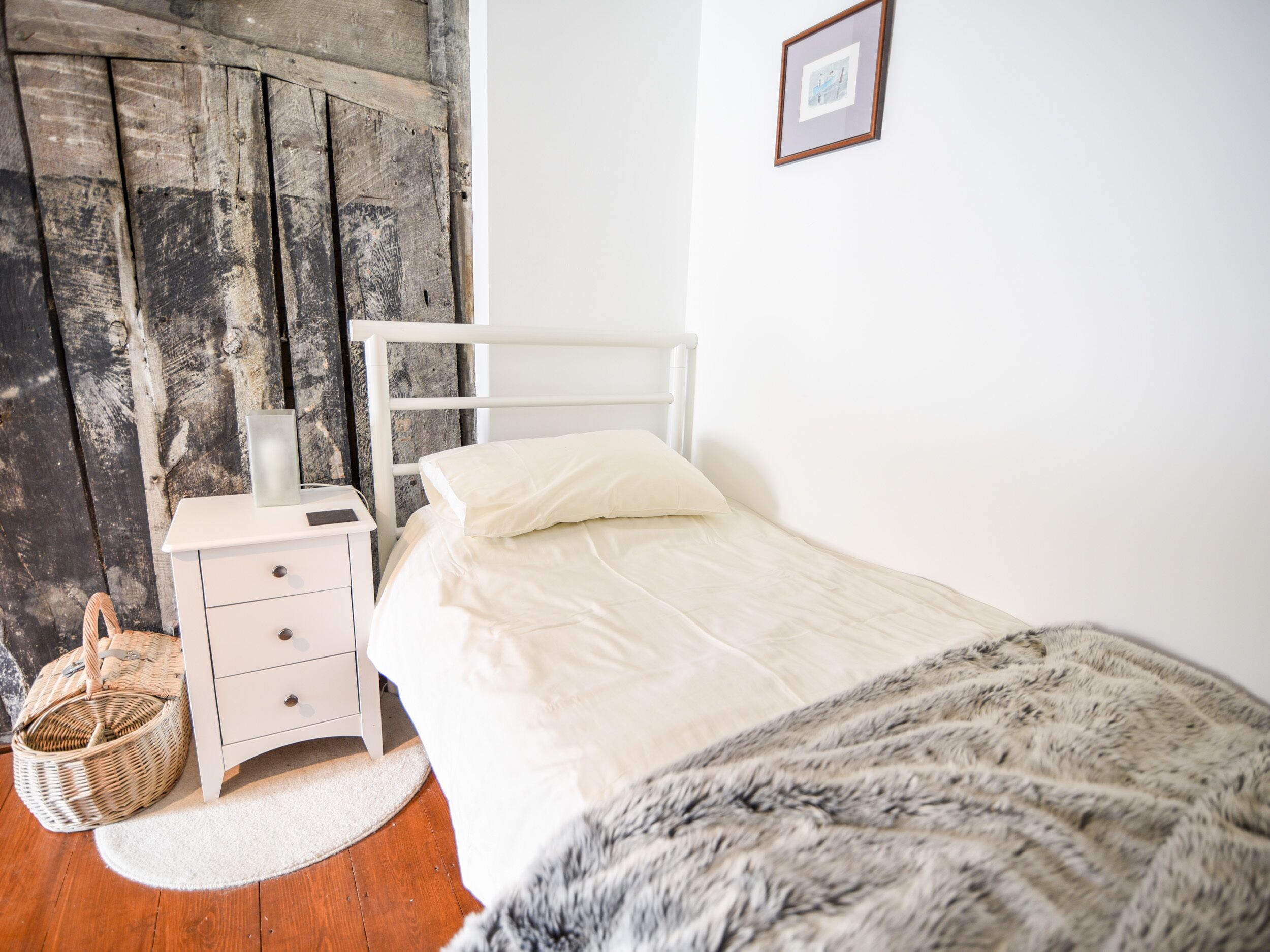 A bedroom with a white bed, a nightstand, a wicker basket, and a wooden door.