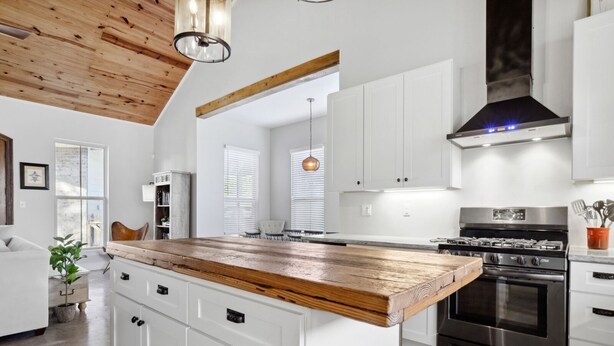 Et moderne køkken med hvide skabe, en træø med stålkant, rustfrie stålapparater og et højt loft med synlige bjælker.