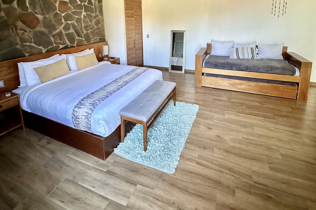 A bedroom with a large bed, a smaller sofa, wooden beams on the ceiling, a stone wall, and an air conditioning unit.