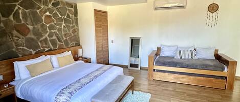 A bedroom with a large bed, a smaller sofa, wooden beams on the ceiling, a stone wall, and an air conditioning unit.