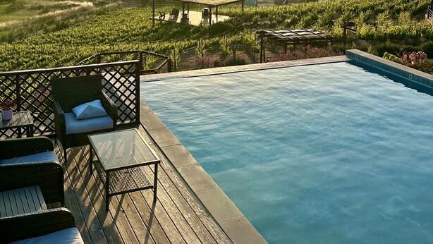 Una piscina con terraza de madera, sillas de descanso y vistas a viñedos y colinas lejanas.