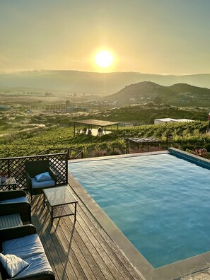 A swimming pool with a wooden deck, lounge chairs, and a view of vineyards and distant hills. - Por Siempre Valle (Valle de Guadalupe)