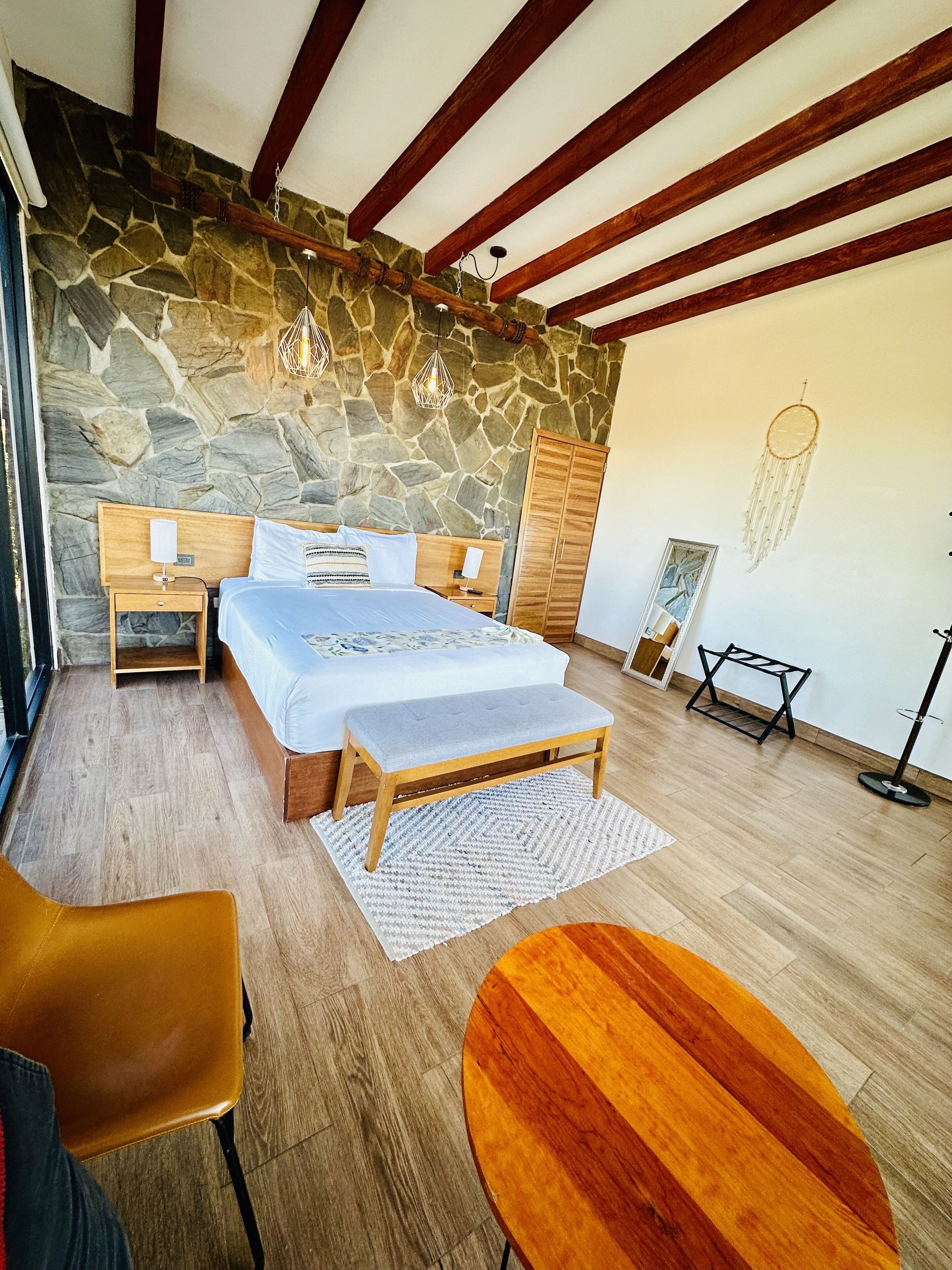 Un dormitorio con pared de piedra, vigas de madera, una cama, mesitas de noche, un banco, una silla, una mesa y un atrapasueños.