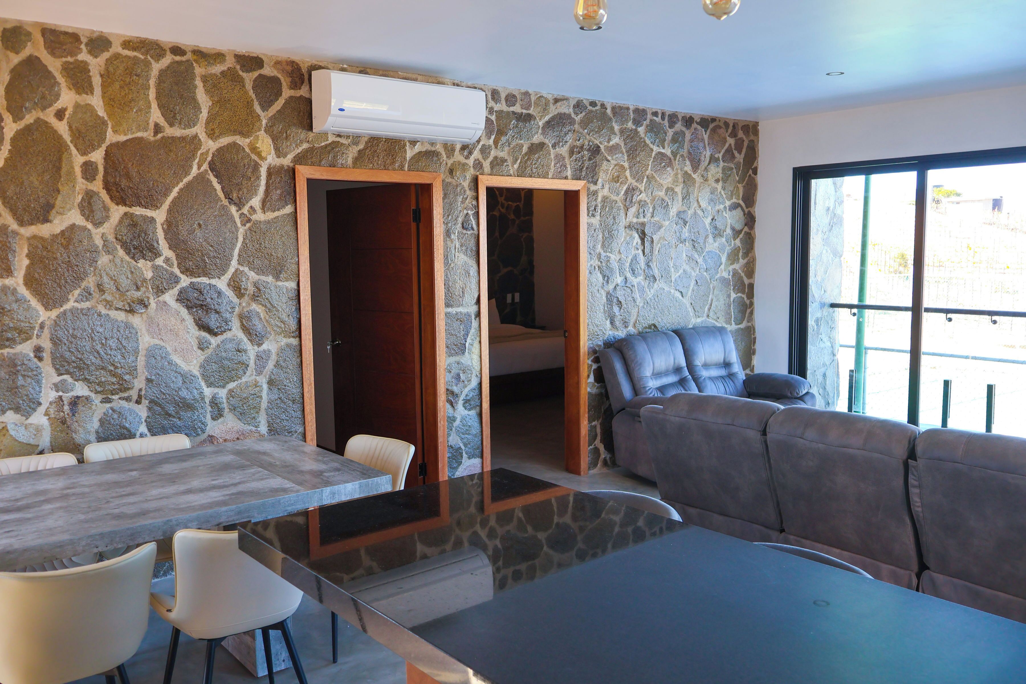 A modern room with a stone wall, a dining table with chairs, a sofa, and a large window.
