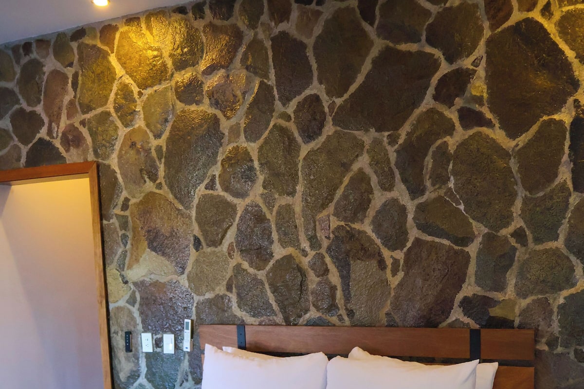 A bed with white linens and a beige throw blanket in a room with a stone wall and wooden headboard.