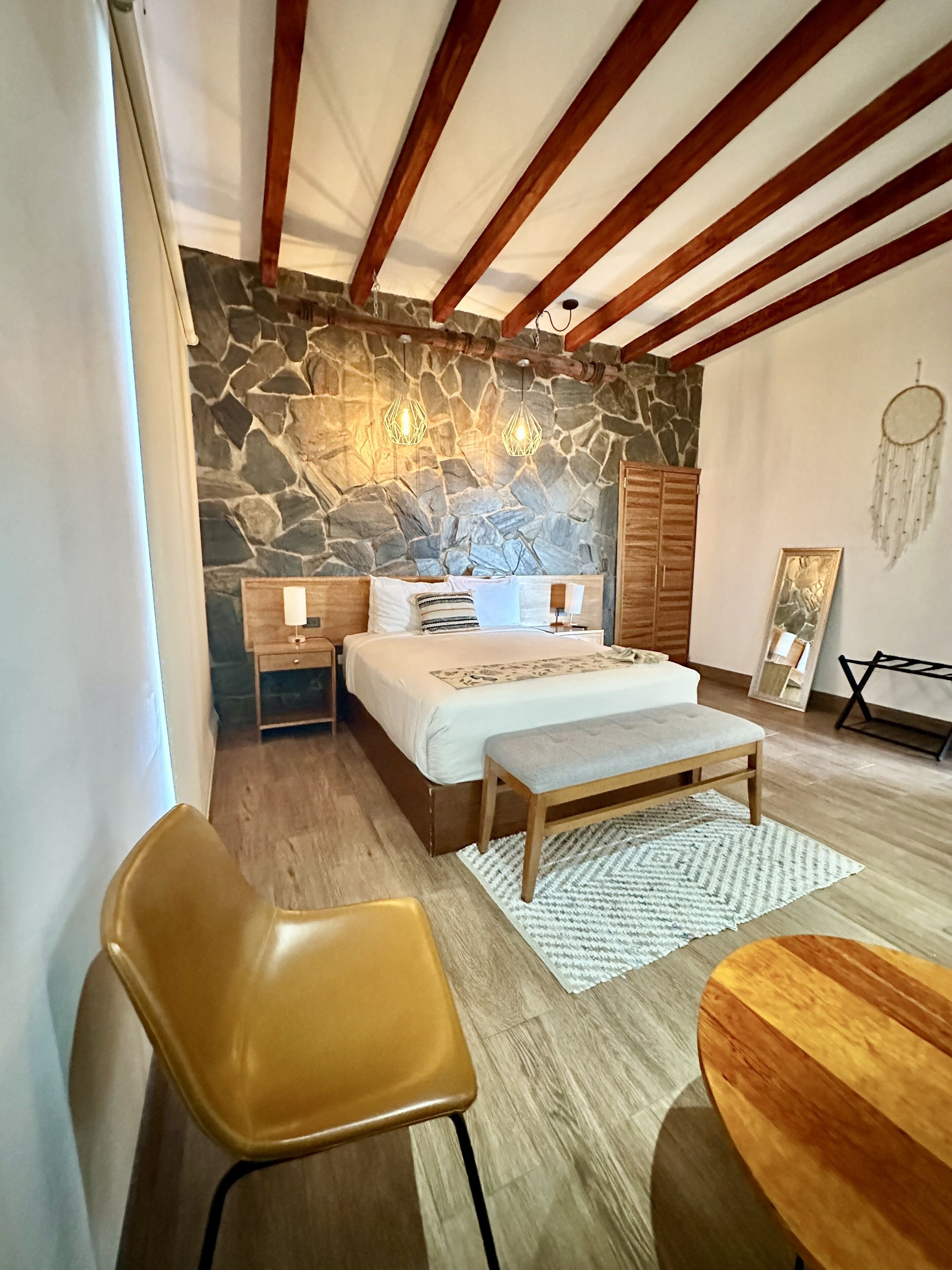 Un dormitorio con pared de piedra, vigas de madera, una cama, mesitas de noche, un banco y un atrapasueños.