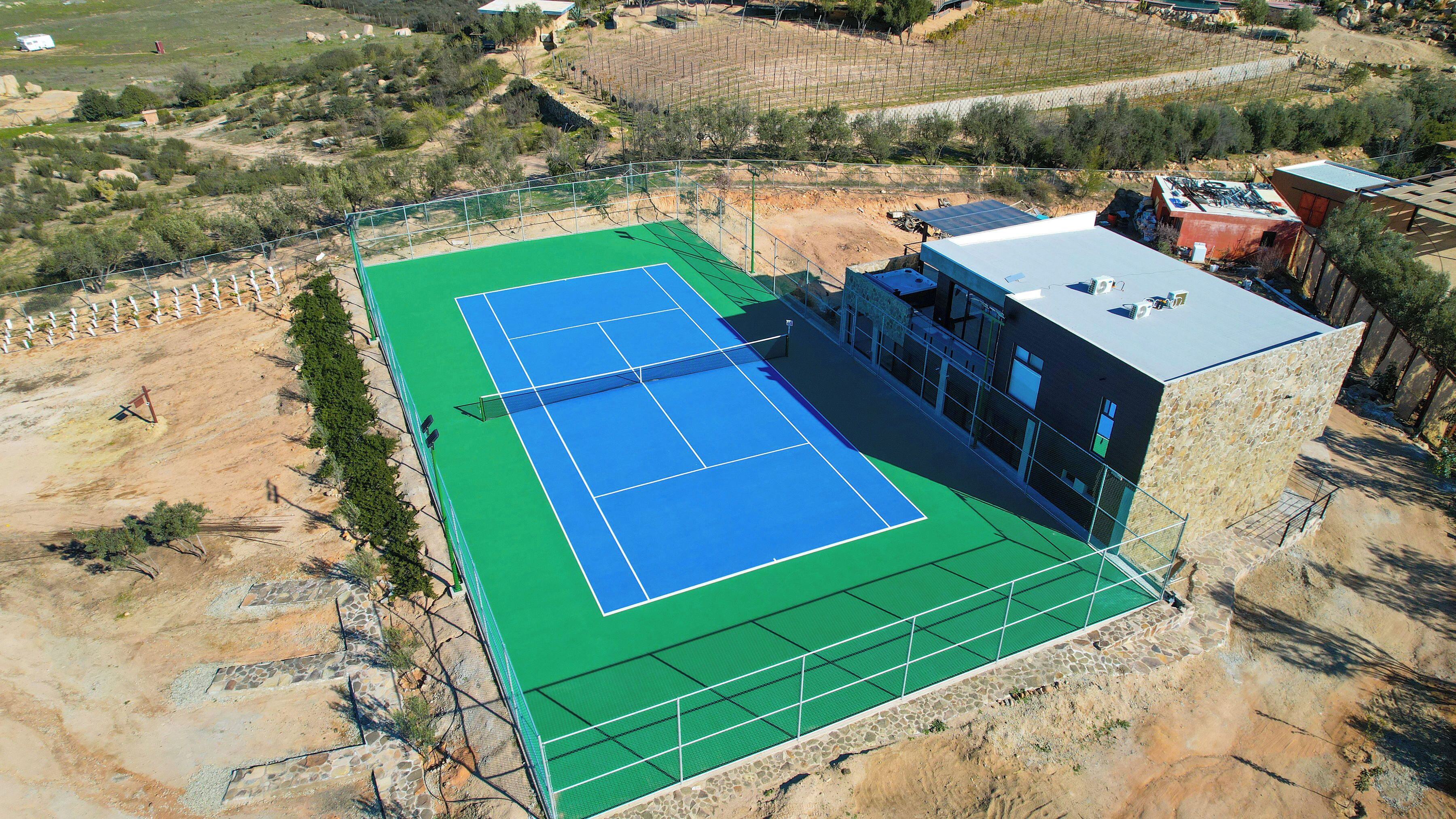 An aerial view of a tennis court with a green and blue surface, surrounded by a fence and a building with a stone facade.