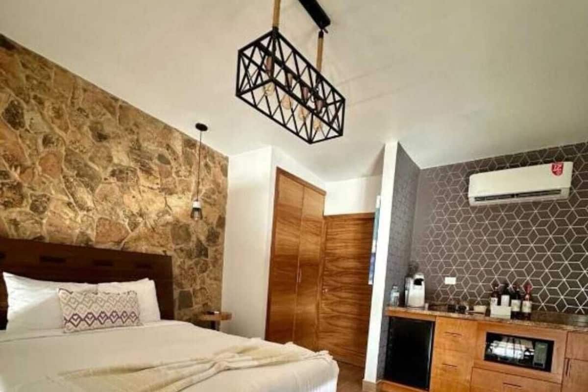 A modern bedroom with a stone wall, a wooden bed, a bench, a wardrobe, and a kitchenette area.