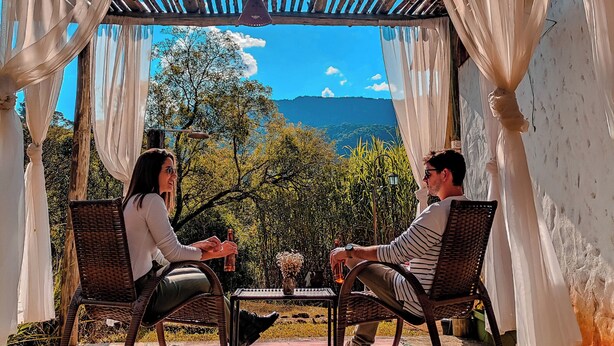 A covered outdoor seating area with two wicker chairs, a small table, and a view of a mountainous landscape. - Serra a vista chalés (Prados)