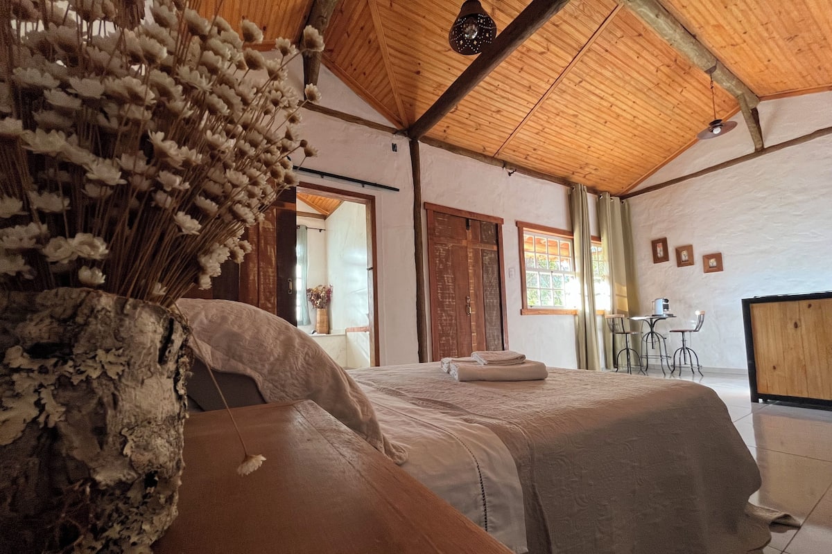 Een kamer met een houten plafond, een bed met een grote decoratieve vaas, een houten bankje, een bureau met een stoel en een raam met gordijnen.