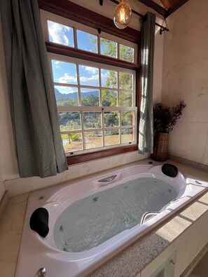 Ein Badezimmer mit Badewanne, einem Fenster mit Vorhängen und Blick auf Grünflächen und Berge.