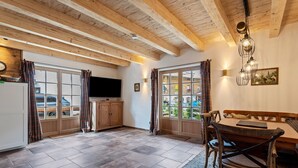 A kitchen with wooden beams, a dining table with chairs, a television, and a view through large windows. - 'Chalet Kaisereck' with Terrace & Wi-Fi (Reit im Winkl)