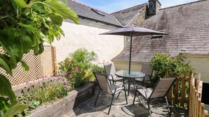 Een terras van natuursteen met een glazen tafel en metalen stoelen, omgeven door een houten hek en planten.