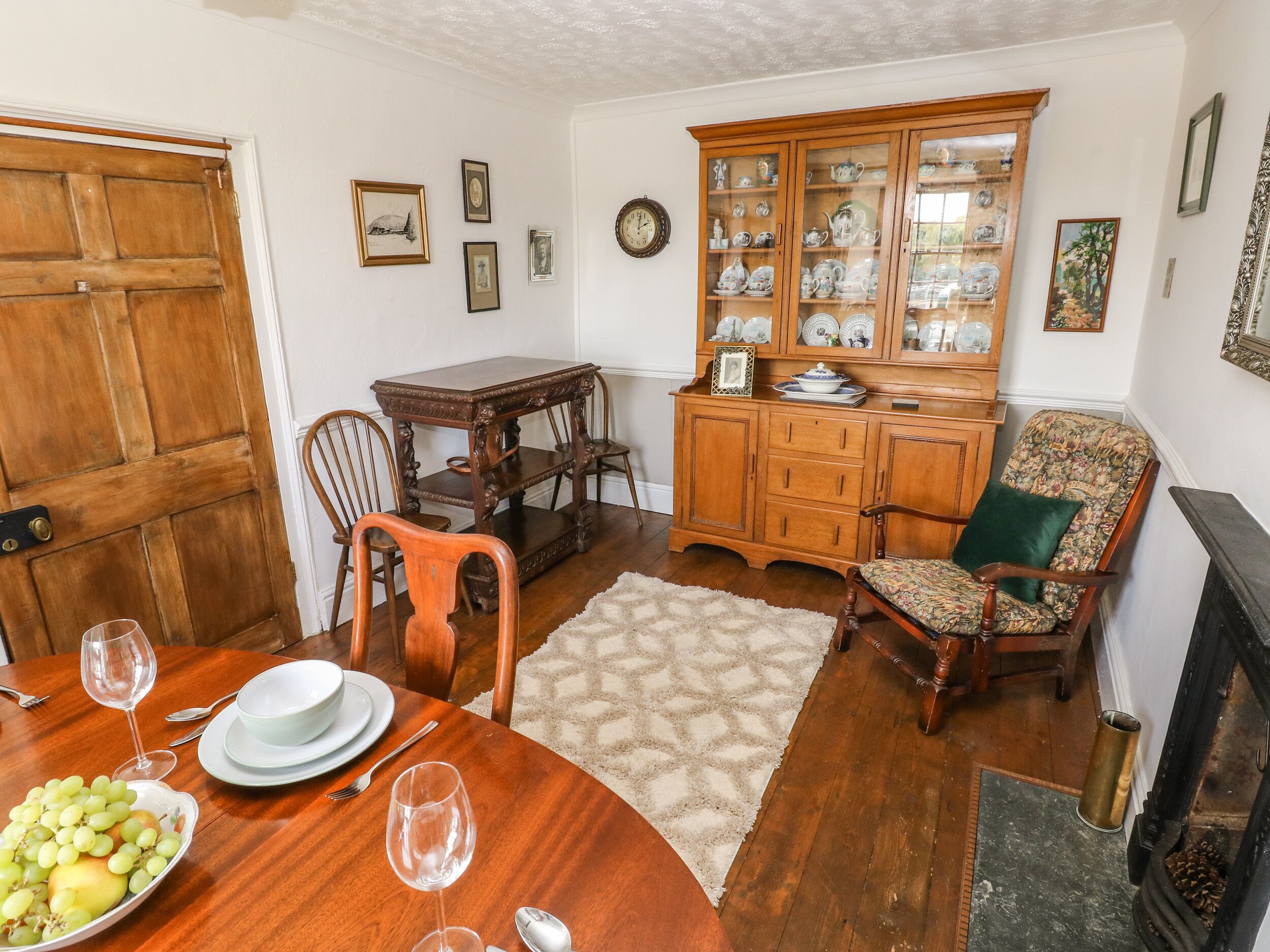 A dining area with a wooden table set for two, a cabinet with glass doors displaying china, and a patterned rug on the floor.