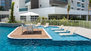 A swimming pool with lounge chairs and a wooden deck area, surrounded by a white fence and modern buildings in the background. - Summit Pinheiros by Okaeri Home Aparts (São Paulo)