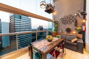 Een moderne woonkamer met een houten eettafel, groene stoelen, een grijze bank en uitzicht op de stad door grote ramen.