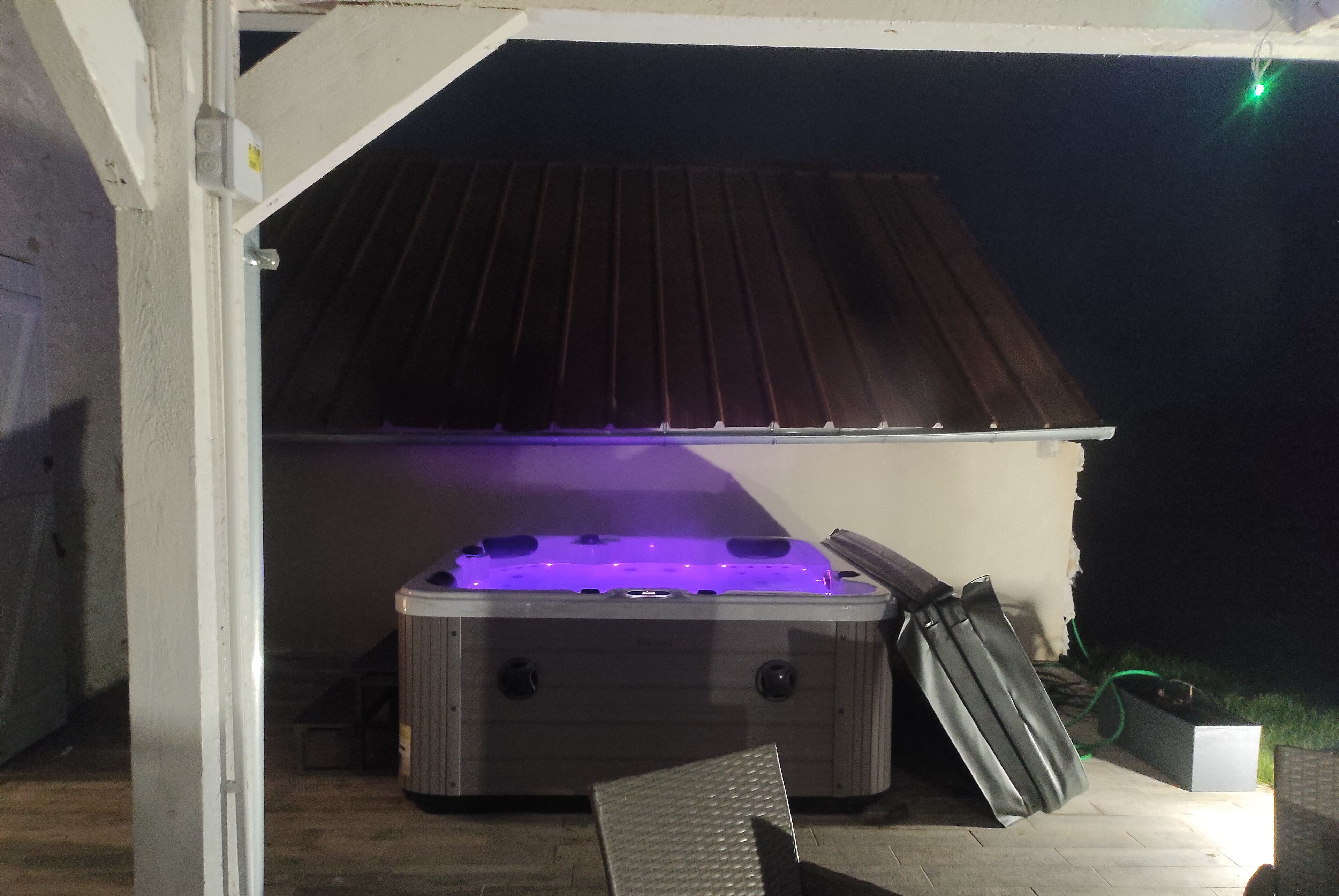 Un jacuzzi bajo un porche cubierto con techo de metal, piso de concreto y una silla metálica.