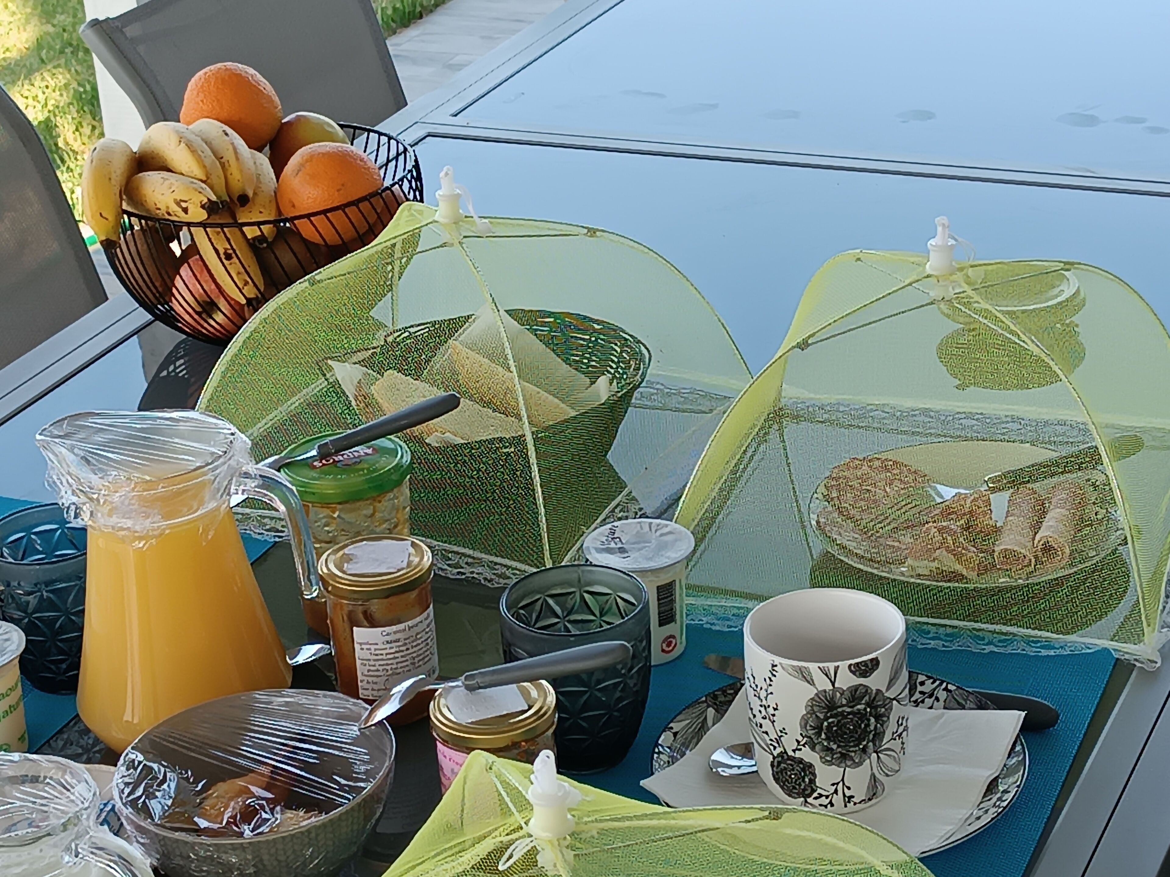 Una mesa con una cesta de frutas, una jarra de jugo, frascos, una taza y un vaso.