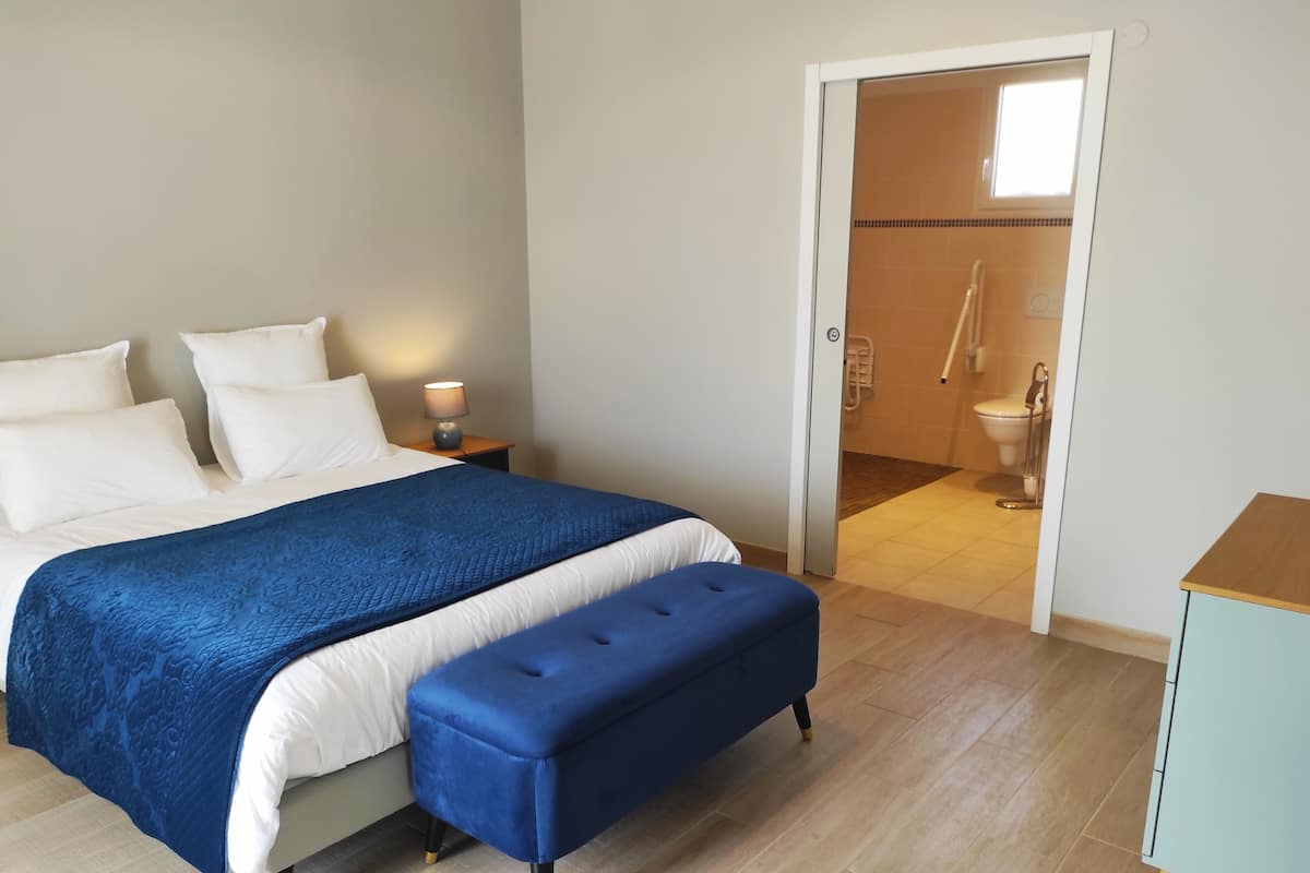 Een slaapkamer met een bed, een nachtkastje, een blauwe bank en een zichtbare badkamer door een open deur.