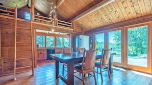 Interior de una cabaña de madera con mesa de comedor, sillas, zona de cocina y ventanas amplias que ofrecen vistas al exterior.