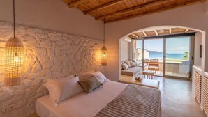 Quarto com parede de pedra, cama, luminária pendente e vista para o mar através de uma porta de vidro.