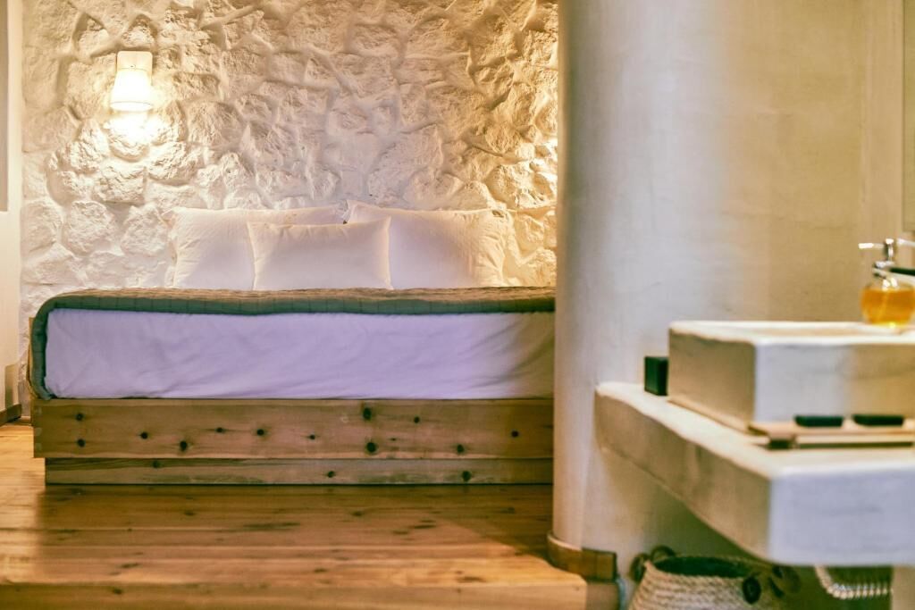 A bedroom with a stone wall, a wooden bed, and a bedside table with a bottle.