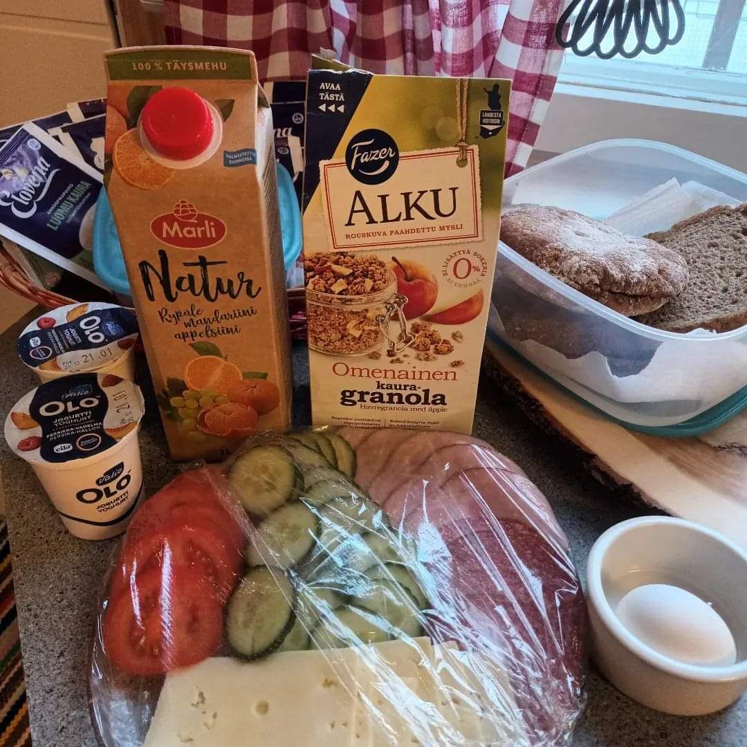 Una encimera de cocina con diversos alimentos, incluyendo un cartón de yogur Marli Natur, un cartón de Alku, un recipiente con granola, una bolsa de plástico con verduras y un bol de huevos.
