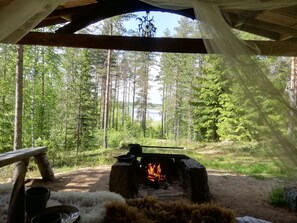 En enkel, rustik träkonstruktion med en eldstad i mitten, omgiven av en skog.