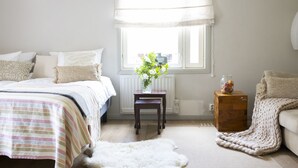 Ein Schlafzimmer mit Bett, Fenster, Heizkörper, einem kleinen Tisch mit Blumenvase und einem Sessel.
