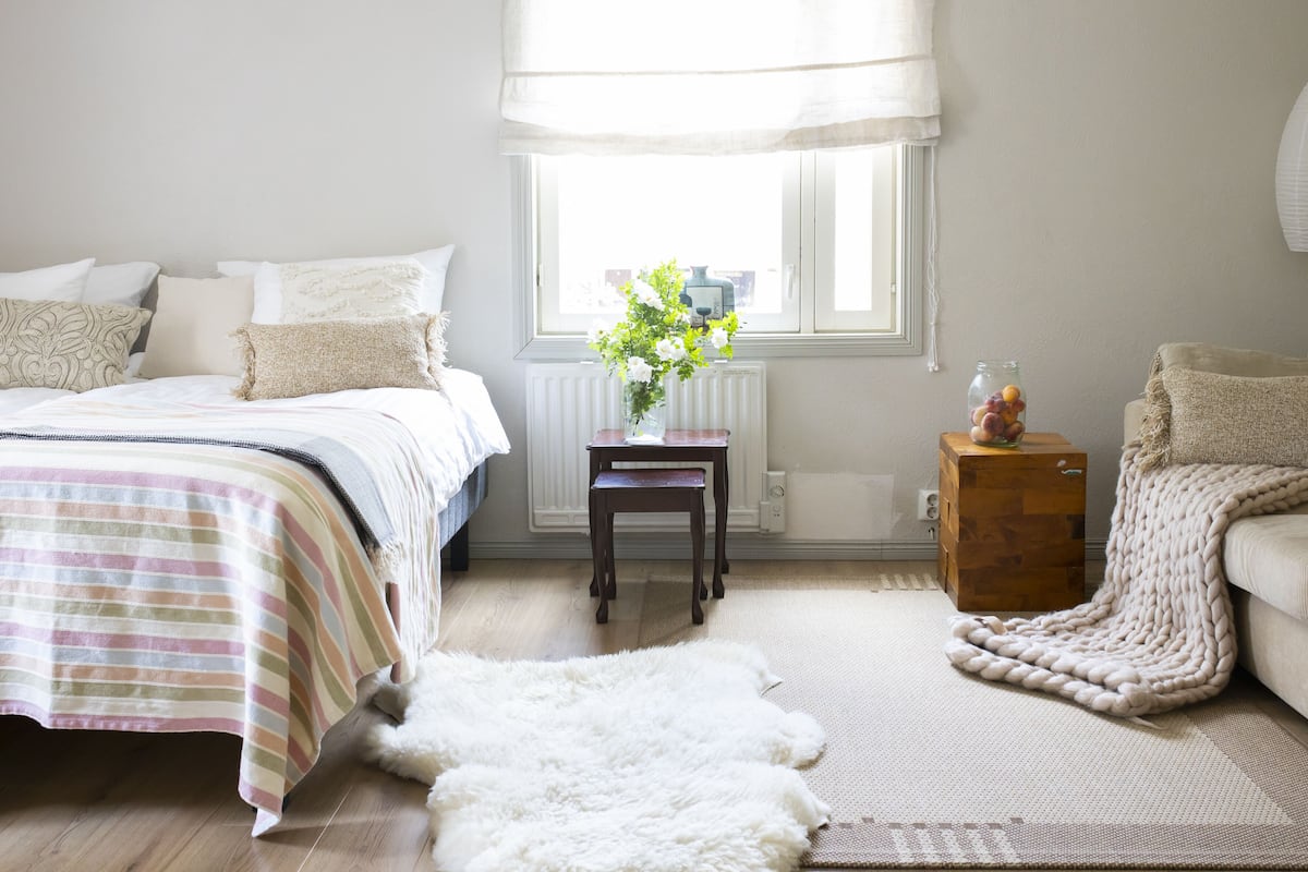 Ein Schlafzimmer mit Bett, Fenster, Heizkörper, einem kleinen Tisch mit Blumenvase und einem Sessel.