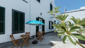 Un edificio blanco con persianas verdes, una sombrilla azul y mobiliario de madera.