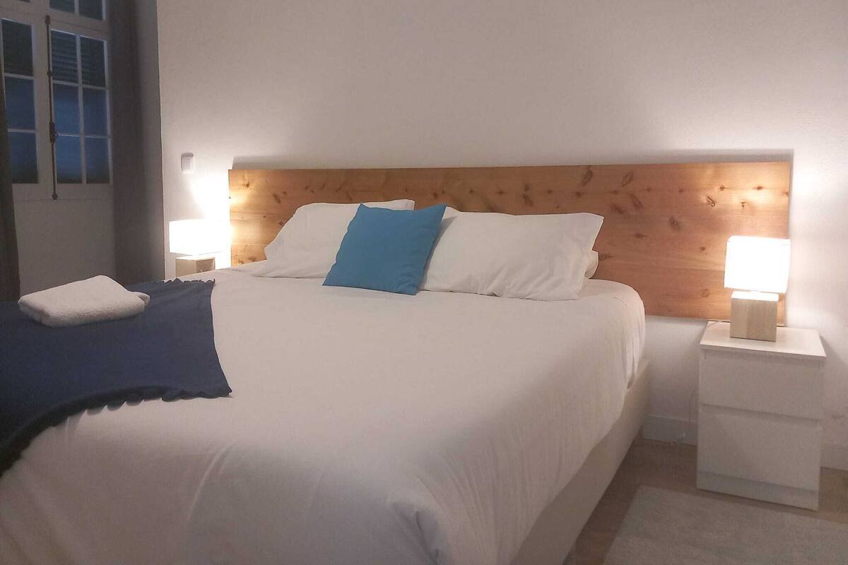 Una cama impecablemente tendida con almohadas blancas y azules, una manta azul y dos lámparas de noche sobre mesitas blancas.