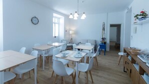 Un comedor moderno y bien iluminado, con mesas y sillas blancas, un reloj en la pared y suelo de madera.