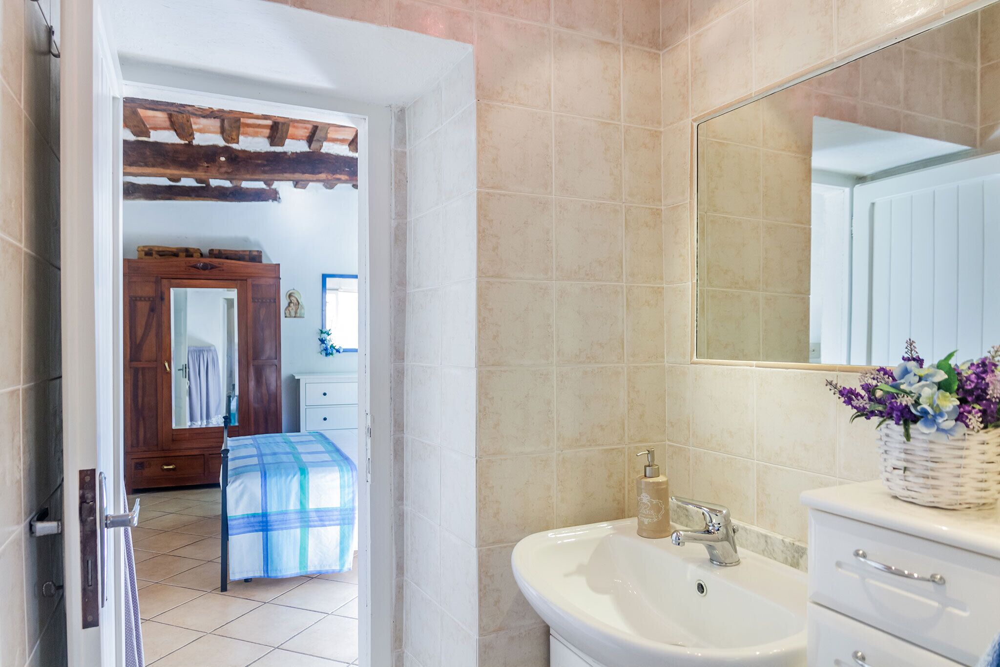 Un baño con un lavamanos blanco, un espejo y una puerta de madera que da a un dormitorio con una cama y una mesita de noche.