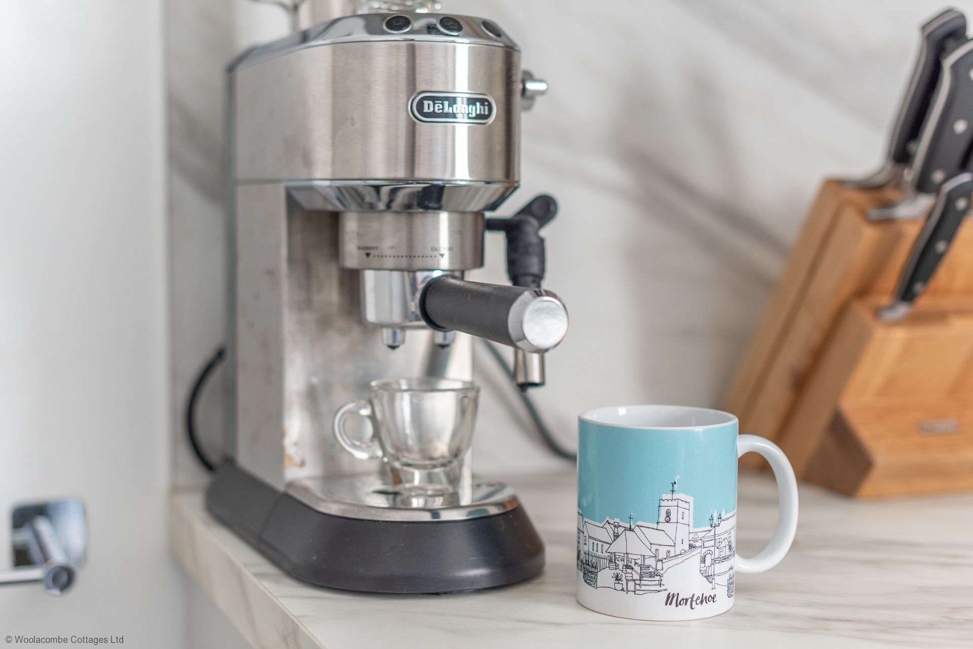 A Delonghi espresso machine with a clear glass cup and a mug with a town illustration.
