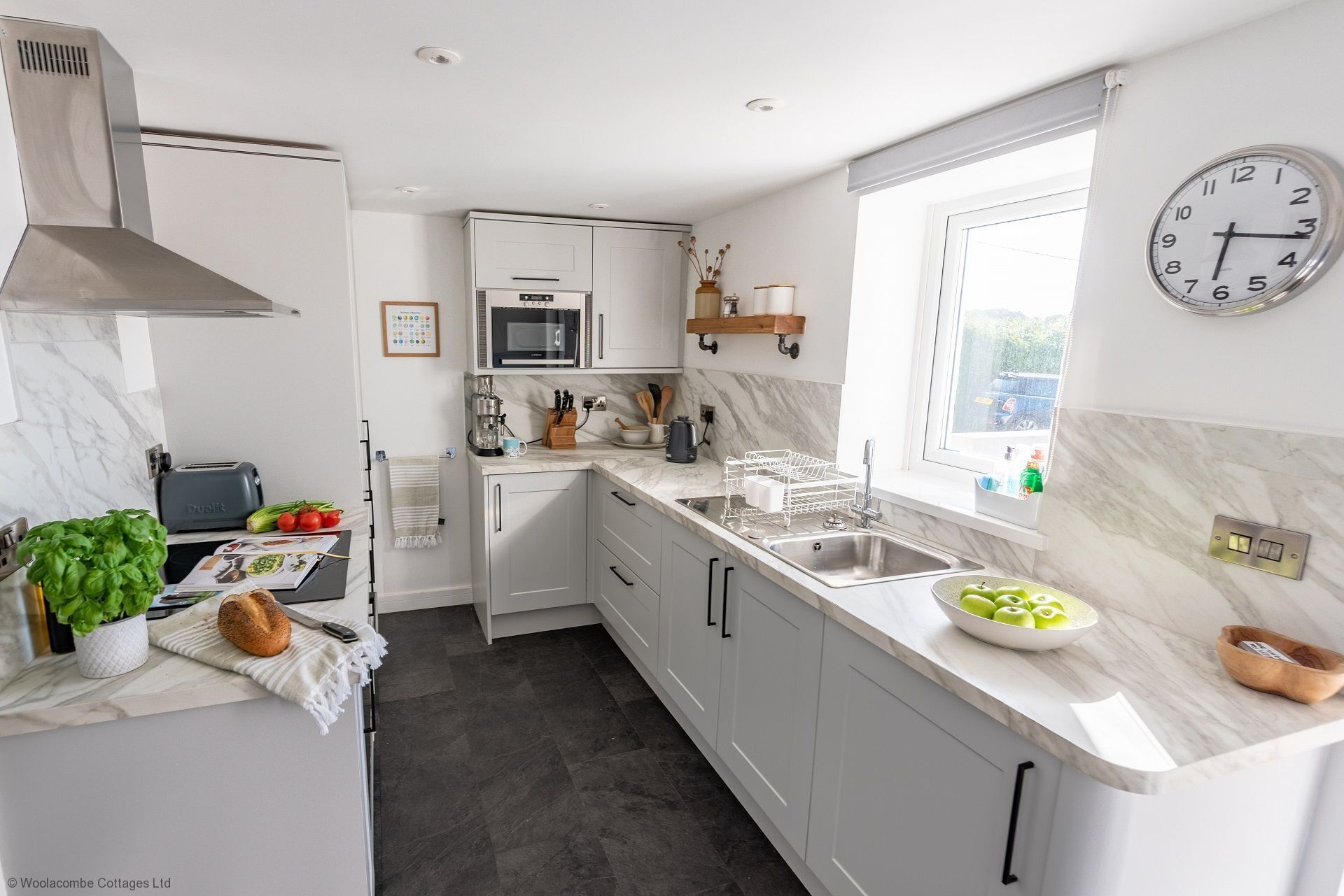 A modern kitchen with white cabinets, a stainless steel sink, a microwave, and a clock on the wall.