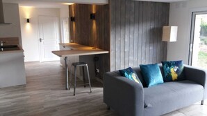 Ett modernt vardagsrum med en grå soffa, en matplats med träbord och ett kök med en bardisk.