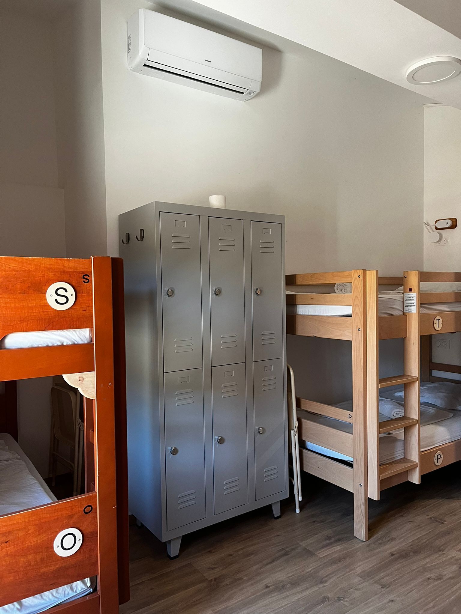 Una cama litera con cabecera de madera, un armario metálico y un equipo de aire acondicionado fijado a la pared.