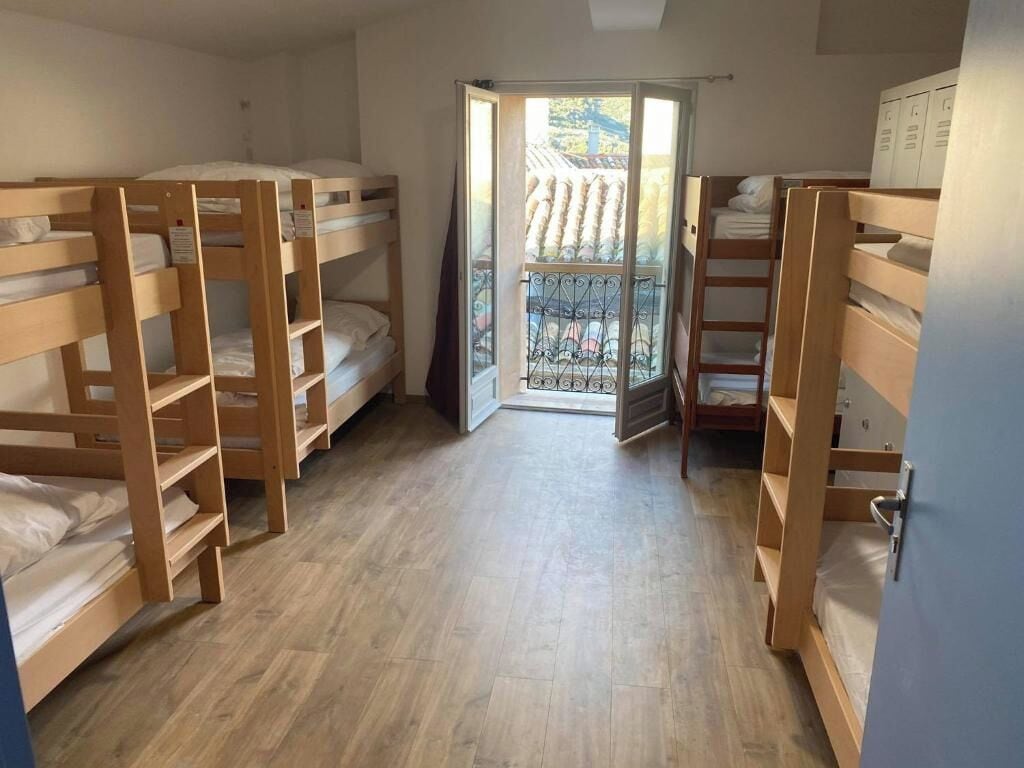 Een slaapzaal met stapelbedden, een balkon met een metalen hekwerk, en een deur die naar buiten leidt.