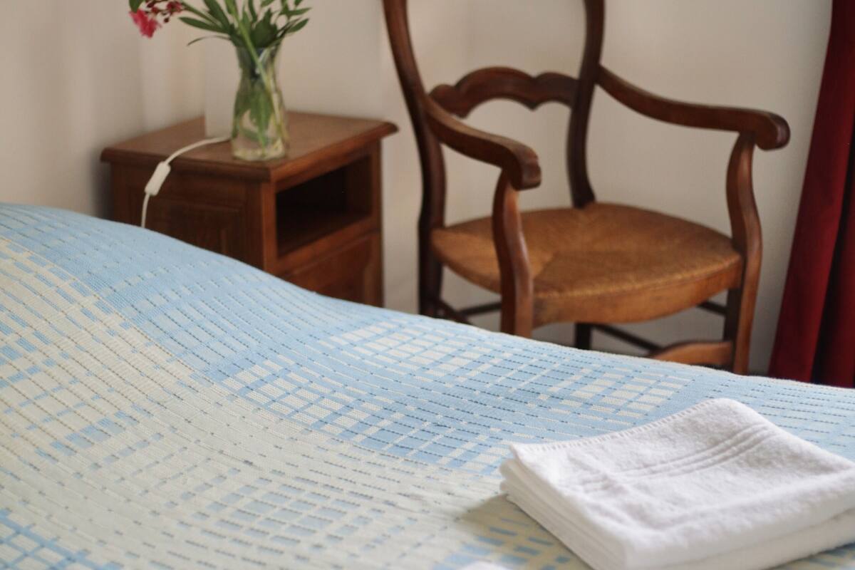 A neatly made bed with a blue and white checkered bedspread, two folded white towels, a wooden nightstand with a vase of flowers, and a wooden chair.