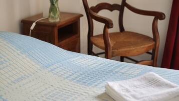 A neatly made bed with a blue and white checkered bedspread, two folded white towels, a wooden nightstand with a vase of flowers, and a wooden chair.