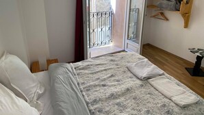 A bedroom with a bed, a wooden floor, a window with a metal gate, and a wooden shelf with clothes.